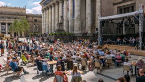 Zdjęcie przedstawia grupę ludzi siedzących na leżakach przed sceną ustawioną w plenerze. Na scenie widzimy muzyków z instrumentami.