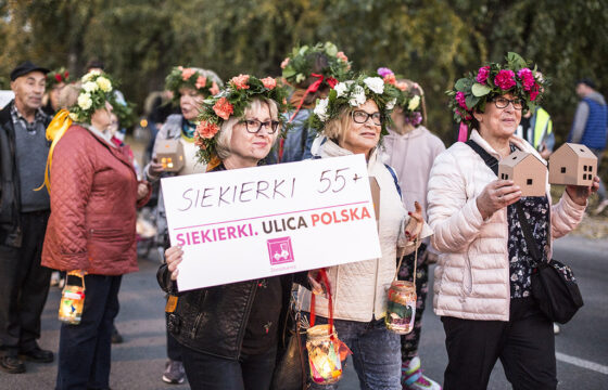 Grupa kobiet na ulicy. Mają wianki z kwiatów na głowie. Jedna trzyma planszę z napisem Siekierki 55+