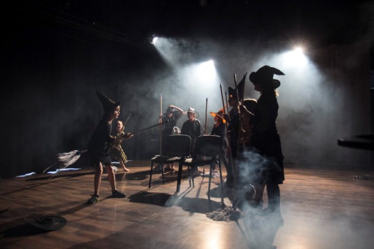 Zdjęcie przedstawia scenę z przdstawienia teatralnego. Na scenie są dziewczynki przebrane za czarownice.