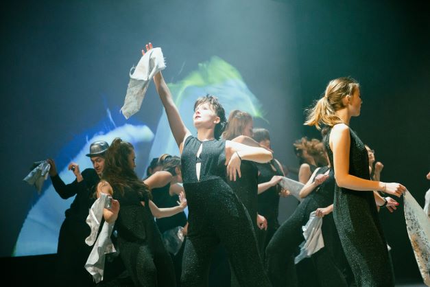 Grupa młodych dziewczyn na scenie, tańczą z chusteczkami w dłoniach, są w jednakowych kostiumach