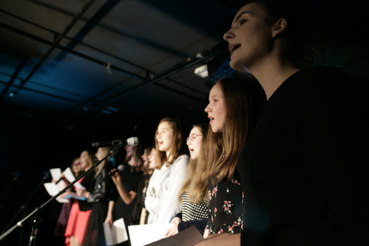 Zdjęcie przedstawia grupę młodzieży na scenie. Stoją przed mikrofonami. Najprawdopodobniej śpiewają.