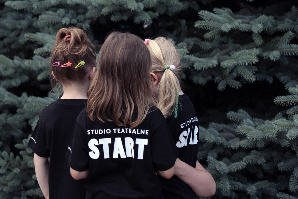 Zdjęcie przedstawia trzy dziewczynki. Są odwrócone plecami do kadru. Mają koszulki z napisem Studio Teatralne Start.
