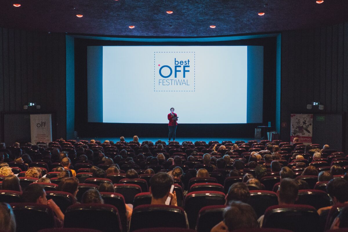 Zdjęcie przedstawia salę kinową. Na ekranie jest logotyp festiwalu Best Off. Na tle ekranu widzimy postać przemawiającą przez mikrofon.