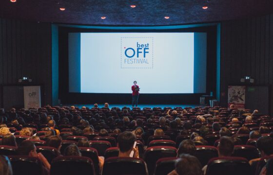 Zdjęcie przedstawia salę kinową. Na ekranie jest logotyp festiwalu Best Off. Na tle ekranu widzimy postać przemawiającą przez mikrofon.