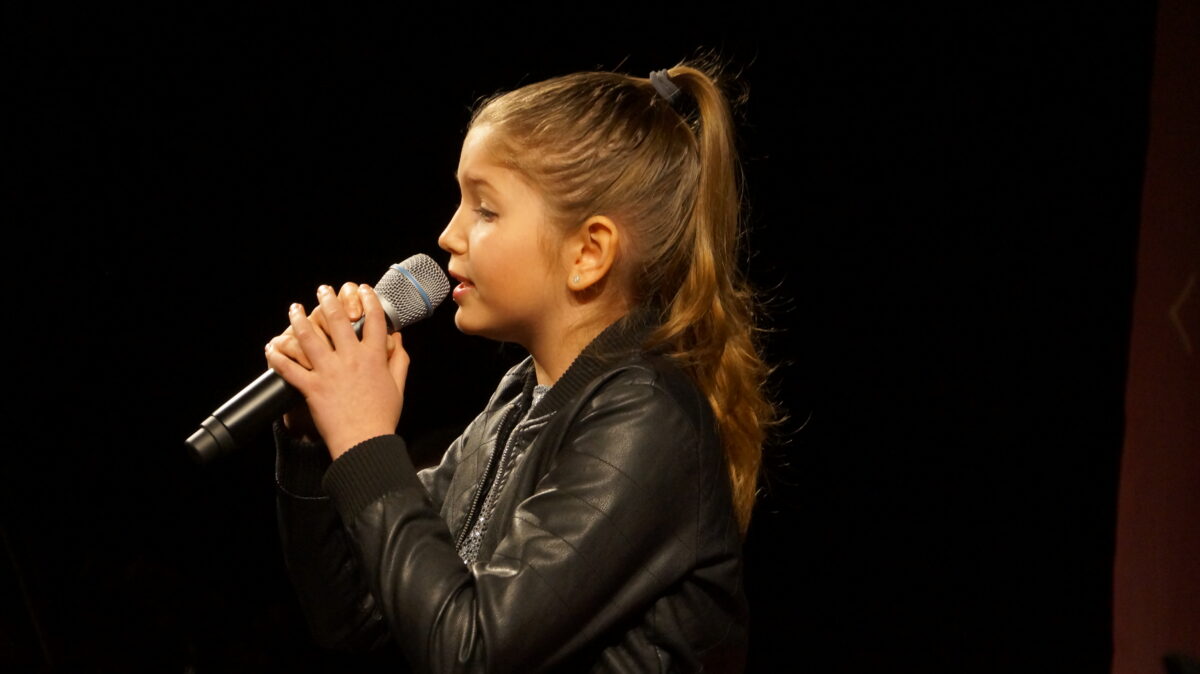 Dziewczynka z mikrofonem w dłoni, uczesana w koński ogon. Zdjęcie zrobione z profilu