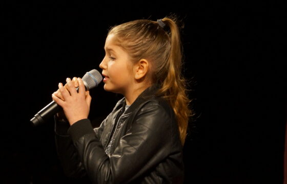Dziewczynka z mikrofonem w dłoni, uczesana w koński ogon. Zdjęcie zrobione z profilu