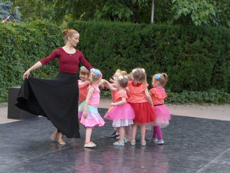 Zdjęcie przedstawia dorosłą kobietę oraz gromadę dzieci w wieku przedszkolnym. Kobieta tańczy, dzieci są z prawej strony kobiety.