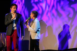 Zdjęcie przedstaawia dwóch chłopców na scenie. W dłoniach mają mikrofony. Ten z lewej strony jest wyższy i wygląda na starszego.