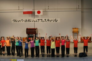 Zdjęcie przedstawia młodzież na dużej hali sportowej. Na ścianie widzą dwie flagi: jedna biało-czerwona i druga biała z czerwonym okręgiem na środku. Pod nimi wisi napis czarnymi japońskimi znakami na białym tle. Młodzież stoi w równym rzędzie na przodzie zdjęcia,wszyscy mają uniesione do góry ręce. Ubrani są w kolorowe, czerwone, zielone, niebieskie, żółte i pomarańczowe koszulki i czarne spodnie.