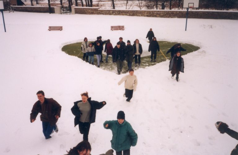 Zima. Grupa młodych ludzi, kilkoro biegnie po śniegu, kilkanaście osób stoi w miejscu odgarniętego śniegu w kształcie serca.