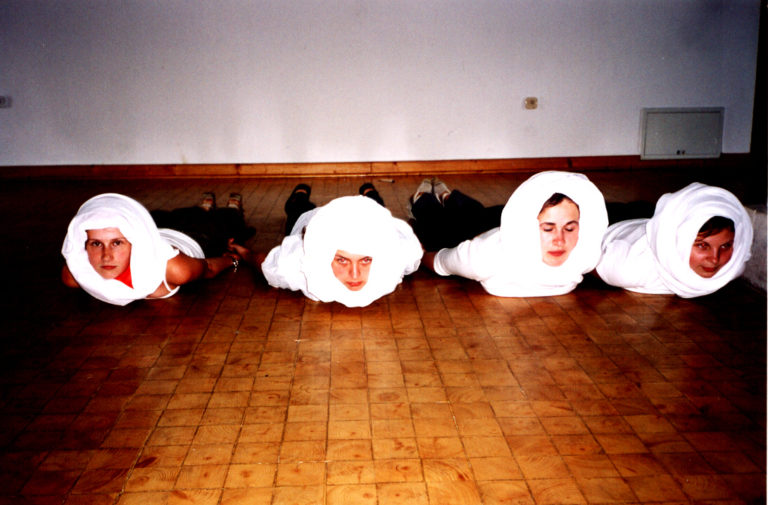 Scena. Na scenie leżą 4 osoby, widać im tylko twarze, głowy mają zawinięte jakimś materiałem.
