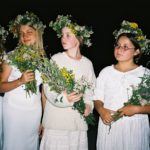 4 dziewczyny, na głowach mają wianki ze świeżych kwiatów, w dłoniach trzymają bukiety kwiatów.