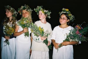 4 dziewczyny, na głowach mają wianki ze świeżych kwiatów, w dłoniach trzymają bukiety kwiatów.