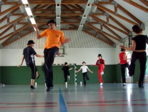 Boisko na sali sportowej, 7 osób tańczy, na głowach mają kapelusze. Ubrani są w spodnie i podkoszulki.