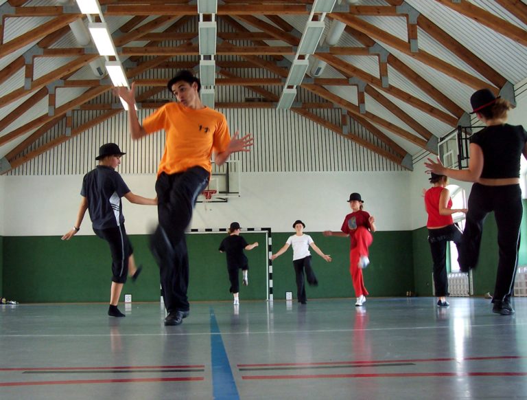 Boisko na sali sportowej, 7 osób tańczy, na głowach mają kapelusze. Ubrani są w spodnie i podkoszulki.