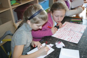 Dwie dziewczynki siedzą przy stole. Jedna z nich pisze na białej kartce, druga opiera się o stół a przed nią leży duża biała kartka zapisana na czerwono.