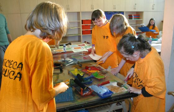 Zdjęcie przedstawia czwórkę dzieci w pomarańczowych koszulkach zgromadzonych wokół stołu z materiałami plastycznymi i malarskimi. Wszyscy patrzą się na rzeczy na stole i malują.