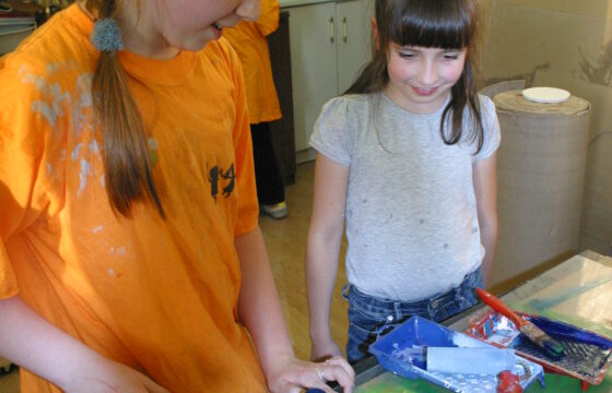 Zdjęcie przedstawia dwie dziewczynki w sali plastycznej. Jedna z nich w pomarańczowej koszulce i warkoczach trzyma w eku wałek umoczony w granatowej farbie. Druga w białej bluzce patrzy się na wałek. Na stole przed nimi leżą artykuły plastyncze i malarskie.