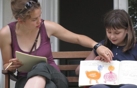 Zdjęcie przedstawia młodą kobietę i dziewczynkę siedzące na ławce. Dziewczynka trzyma przed sobą otwarty notes z rysunkami przedsawiającymi ptaki. Kobieta pokazuje ręką na rysunki.