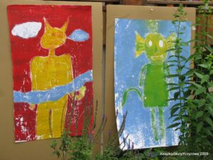 Zdjęcie przedstawia dwie prace plastyczne powiedzone w galerii na płocie. Praca po lewej stronie przedstawia żółtego kota na czerwonym tle, a praca po prawej przedstawia zielonego stworka z dużymi uszami na niebieskim tle.