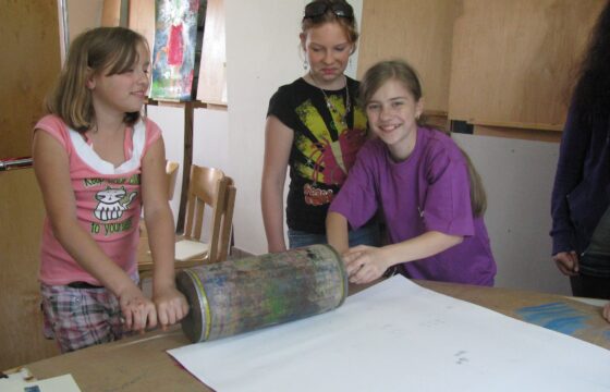 Trzy dziewczynki stoją za dużym stołem, na którym leży duża biała kartka papieru. Dwie dziewczynki trzymają z dwóch stron duży wałek