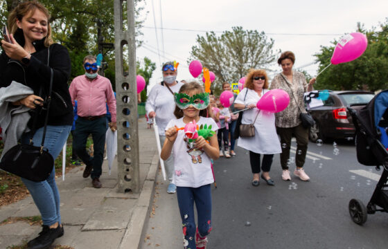 Zdjęcie przedstawia kolorowy tłum idący ulicą. Na środku idzie mała dziewczynka w białej bluzce i zielonej masce karnawałowej na twarzy. Za nią idą kobiety i mężczyźni trzymający różowe balony.
