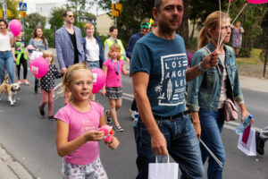 Grupa kolorowo ubranych osób idzie ulicą. Na pierwszym planie jest dziewczynka, mężczyzna trzymający różowe balony i kobieta trzymająca kolorową maskę.
