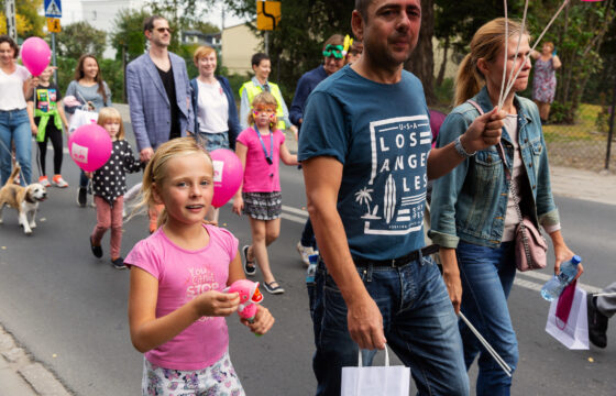 Grupa kolorowo ubranych osób idzie ulicą. Na pierwszym planie jest dziewczynka, mężczyzna trzymający różowe balony i kobieta trzymająca kolorową maskę.