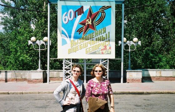 Dwie kobiety w okularach przeciwsłonecznych stoją przed plakatem z napisem 60 i hasłem w cyrylicy. Nad plakatem powiewają dwie czerwone flagi.