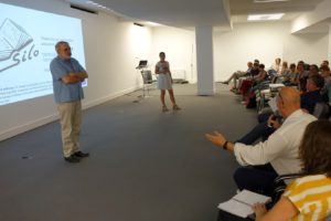 Spotkanie osób. Na scenie kobieta i mężczyzna. Przed nimi grupa osób siedzących na krzesłach.