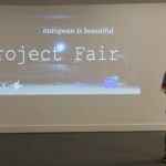 Mężczyzna stoi na scenie. Za nim na ekranie wyświetla się napis" Project fair. European is beautiful"