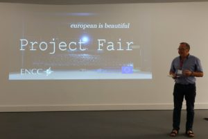 Mężczyzna stoi na scenie. Za nim na ekranie wyświetla się napis" Project fair. European is beautiful"