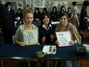 Zdjęcie przedstawia trzy młode kobiety. Dwie z nich o europejskich rysach twarzy trzymają w rękach białe kartki papieru z kolorowymi brzegami, na których zapisane są japońskie znaki. Po środku siedzi młoda uśmiechająca się dziewczyna o azjatyckich rysach twarzy.