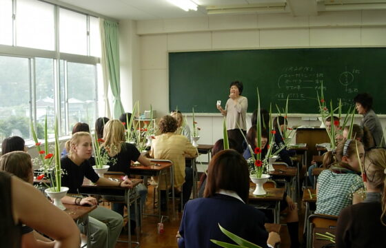 Zdjęcie przedstawia klasę szkolną w Japonii. Po lewej stronie jest ściana okien, w tle biała ściana z zieloną tablicą kredową. Przy ławkach tyłem siedzi młodzież. Na stołach stoją rośliny i kwiaty w białych ceramicznych wazonach. Na środku w głębi widać kobietę o azjatyckich rysach twarzy tłumaczącą coś dzieciom.