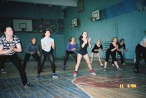Zdjęcie przedstawia zajęcia taneczne na sali gimnastycznej. Grupa siedmiu dziewcząt stoi w rozkroku opierając ręce na kolanach. Przed nimi, tyłem do aparatu, stoi kobieta pokazująca pozę.