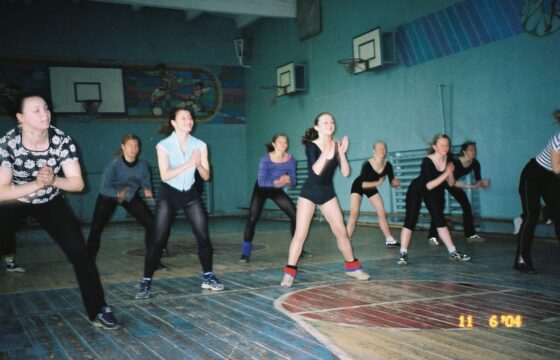 Zdjęcie przedstawia zajęcia taneczne na sali gimnastycznej. Grupa siedmiu dziewcząt stoi w rozkroku opierając ręce na kolanach. Przed nimi, tyłem do aparatu, stoi kobieta pokazująca pozę.