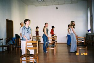 Zdjęcie przedstawia zajęcia taneczne w sali szkolnej. Grupa dziewcząt stoi bokiem każda przy swoim krześle, trzymając na nim jedną rękę