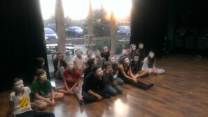 Zdjęcie przedstawia grupę ok 20 młodych osób w białych maskach na twarzy. Maski nie mają żadnego wyrazu, są neutralne. Młodzież siedzi na drewnianej podłodze, za nimi jest ściana z okien, za oknem widać parking i drzewa