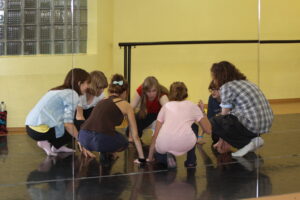 Zdjęcie przedstawia grupę dziewcząt kucających w kręgu na sali tanecznej.
