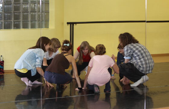 Zdjęcie przedstawia grupę dziewcząt kucających w kręgu na sali tanecznej.