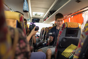 Zdjęcie przedstawia wnętrze autokaru. Na siedzeniach siedzą młode osoby, jedna z nich trzyma aparat fotograficzny w ręku.