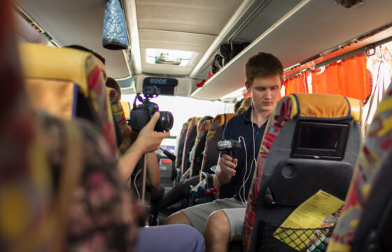 Zdjęcie przedstawia wnętrze autokaru. Na siedzeniach siedzą młode osoby, jedna z nich trzyma aparat fotograficzny w ręku.