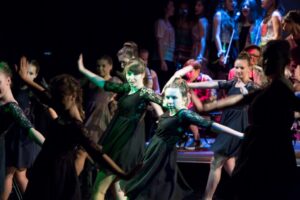 Zdjęcie przedstawia fragment spektaklu. Grupa dziewcząt w zcarnych sukienkach z koronkowymi rękawami stoi w tanecznej pozie - twarze są zwrócone do aparatu, ciała lekko w lewą stronę, ręce wyciągnięte na boki.