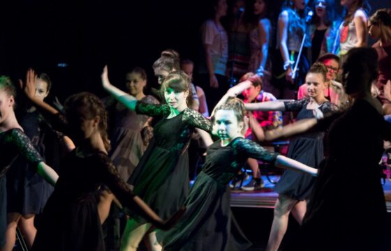 Zdjęcie przedstawia fragment spektaklu. Grupa dziewcząt w zcarnych sukienkach z koronkowymi rękawami stoi w tanecznej pozie - twarze są zwrócone do aparatu, ciała lekko w lewą stronę, ręce wyciągnięte na boki.