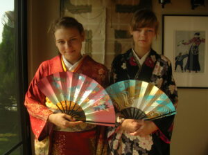 Zdjęcie przedstawia dwie dziewczyny w tradycyjnych, wzorzystych i kolorowych kimonach. Dziewczynki trzymają w rękach kolorowe wachlarze.