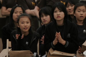 Zdjęcie przedstawia grupę klaszczących dziewcząt. Widać je od pasa w górę, ubrane są w czarne stroje z żółto-złotym pasem. Dziewczęta mają długie czarne włosy i azjatyckie rysy twarzy.
