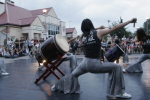 Zdjęcie przedstawia fragment koncertu na bębnach. Ujęcie pokazuje grupę osób dynamicznie grających na dużych bębnach wspartych na drewnianych stojakach. Osoby grające są tyłem, stoją szerokim rozkroku i mają wyciągniętą ręke, w której trzymają pałki do gry. Ubrane są w cekinowe srebrne spodnie i czarne koszulki z białym napisem po japońsku na plecach. W tle widać widownię siedzącą na krzesłach i budynek z czerwonym spadzistym dachem