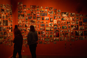 Zdjęcie przedstawia czerwoną dużą ścianę, na której przyklejone są zdjęcia. Pod ścianą widać ciemne sylwetki dwóch stojących osób.