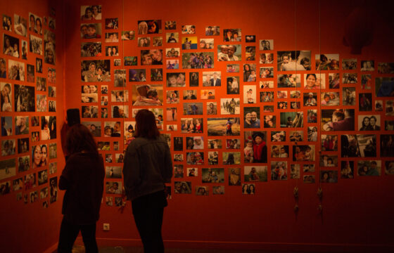 Zdjęcie przedstawia czerwoną dużą ścianę, na której przyklejone są zdjęcia. Pod ścianą widać ciemne sylwetki dwóch stojących osób.