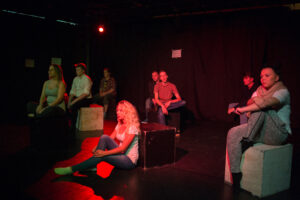 Zdjęcie przedstawia scenę ze spektaklu. Grupa młodych aktorów siedzi na czarnych kubikach, oświetlenie jest czerwone.